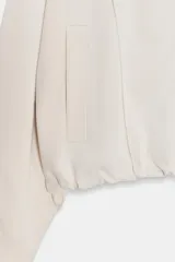 Cazadora estilo bomber de color beige, con cuello subido, manga larga y cierre frontal con cremallera. Presenta bolsillos delanteros de vivo y bajo ajustable con cordones elásticos laterales.