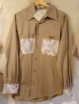 Camisa beige de manga larga, con bolsillos frontales en el pecho, puños y solapas de los bolsillos con estampado floral y ornamental.