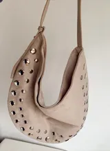 Cartera tipo hobo de símil gamuza color beige, con diseño de media luna y decorada con tachas metálicas plateadas en todo su contorno. Incluye correa ajustable y bolsillo interno.