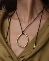 Collar con dije colgante de forma orgánica e irregular, acabado dorado, suspendido de un cordón negro de algodón natural.