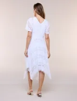 Vestido midi blanco de estilo romántico confeccionado en poliéster con elastano, con detalles de encaje floral. Cuenta con escote cruzado en V, mangas cortas acampanadas, cintura elastizada y falda evasé con terminación asimétrica en picos.