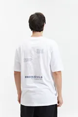 Remera blanca de manga corta con ajuste regular. Presenta un estampado en la espalda con un diseño floral lineal en gris, texto en inglés y el logo "BROOKSFIELD The vacation era" en azul.