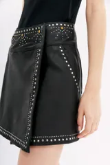 Falda corta negra de cuero ovino con diseño cruzado tipo wrap. Presenta apliques de tachas y piedras plateadas y doradas a lo largo de los bordes y en la cintura.