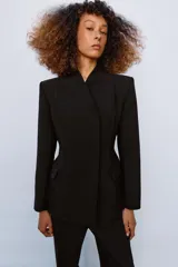 Blazer entallada de color negro con escote en pico, manga larga con hombreras y cierre frontal con botones ocultos. Presenta bolsillos con solapa en el delantero y detalle de nervios marcados en el tejido.