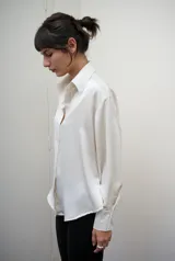 Camisa de manga larga confeccionada en satén de color blanco, con superficie lisa, leve brillo y caída suave. Presenta cuello clásico y cartera frontal.