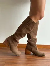 Botas de caña alta confeccionadas en cuero gamuzado color marrón. Presentan un diseño de estilo western con puntera cuadrada, taco de madera y una tira decorativa con estribo metálico desmontable en la zona del tobillo.