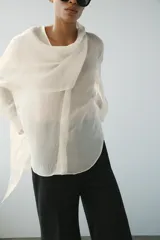 Camisa de hilatura con mezcla de lyocell color blanco, con cuello fluido con pañuelo incorporado, manga larga con puño y bajo redondeado asimétrico. Cierre frontal con botones ocultos.