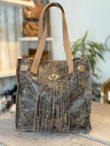 Cartera tipo tote de cuero color marrón con efecto craquelado y flecos. Tiene doble asa de hombro color beige y cierre de gancho metálico.