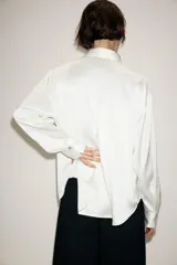 Camisa de satén color blanco, de corte clásico con cuello solapa, manga larga con puños y cierre frontal con botones dorados.