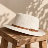 Sombrero de paja color blanco, estilo fedora, con cinta marrón de cuerina y hebilla metálica dorada decorativa en el lateral.