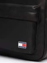Mochila negra de poliuretano con acabado liso y diseño curvo. Tiene un compartimento principal con cierre de cremallera oculta, asa superior, correas ajustables y acolchadas, bolsillo interior de malla y bolsillo exterior con cremallera. Incluye inscripción de Tommy Jeans en las tiras e insignia de Tommy en la parte delantera.