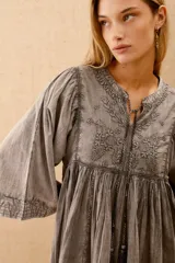 Vestido corto de algodón color gris lavado, con escote en V y cordones ajustables. Presenta mangas anchas tipo murciélago y bordados florales en el canesú.