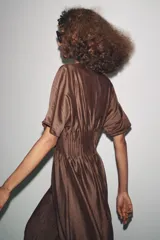 Vestido midi color marrón con textura brillante y arrugada, cuello redondo y mangas cortas tipo murciélago. Presenta un canesú fruncido con pliegues verticales en la cintura que da paso a una falda de corte A.