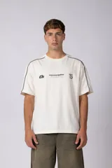 Remera de algodón color blanco con corte regular, cuello redondo y mangas cortas. Presenta detalles de líneas finas negras en los hombros y estampado gráfico frontal con tipografía y logotipos en color negro.