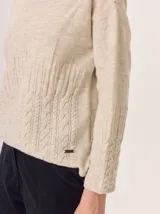 Buzo de lana color beige claro, con cuello chimenea y manga larga. Presenta trenzas de punto grueso en el frente y en los puños, con un diseño texturizado que se asemeja a espigas o ramas.