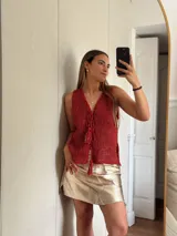 Chaleco tejido en punto calado con hilo de algodón y lurex, de color negro con destellos plateados. Presenta escote profundo en V, cierre frontal con cordones entrelazados terminados en borlas y un corte recto y holgado.