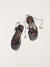 Sandalias planas de cuero negro estilo romano, con tiras finas cruzadas, detalle de anillo central con cordones decorativos y cuentas de colores en los extremos de las tiras.