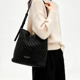 Cartera de hombro Korium modelo Alina, de diseño trenzado en color negro, con correa regulable y aplique metálico con el logo de la marca.