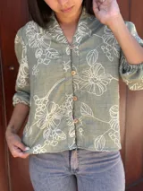 Camisa de corte holgado en color verde con estampado floral lineal en tono crema. Presenta cuello mao, cierre frontal con botones y mangas tres cuartos con puños elásticos.