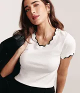 Blusa de algodón acanalado blanco, de corte ajustado, con mangas cortas y cuello redondo. Presenta un delicado ribete ondulado en contraste de color negro en el cuello y en los puños de las mangas.