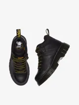 Bota de montaña Dr. Martens modelo Woodard, color negro, con cordones de tipo excursionista en amarillo y negro, cuello acolchado, suela acanalada Club Wedge con pespunte negro y tirador de talón de goma.