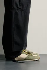 Zapatilla deportiva barefoot fabricada en combinación de piel y tejido, en tonos verde oliva y beige, con cordones elásticos amarillos y suela de goma flexible.