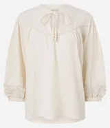 Blusa estilo bata color blanco roto, confeccionada en cambraia de algodón y lino. Presenta cuello con lazo ajustable, canesú fruncido en el busto y mangas tres cuartos con puños elásticos y bordados.