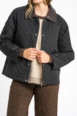 Campera negra de diseño capitoneado con cuello de solapa en contraste color marrón. Presenta cierre frontal con cremallera y botones a presión, mangas largas con puños ajustables y un corte recto.