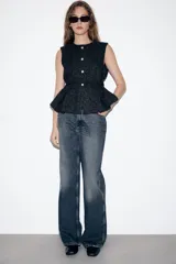 Chaleco de tejido tipo tweed en color negro, con cuello redondo y cierre frontal de botones dorados. Presenta un diseño entallado con cinturón integrado del mismo tejido y bajo con pliegues.