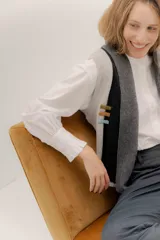 Chaleco abierto de tejido de punto, con diseño de bloques de color verticales en gris claro, negro y gris oscuro. Presenta un cuello ancho tipo chal y un detalle de tres pequeñas aplicaciones bordadas horizontales en tonos verde oliva, naranja quemado y celeste pálido en el frente.