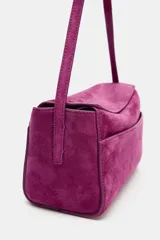 Mini bolso de mano confeccionado en piel de gamuza color fucsia. Presenta un diseño estructurado con asa corta, cierre principal mediante cremallera y solapa, y bolsillos exteriores en la parte frontal y trasera.