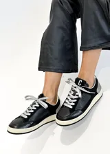Zapatillas urbanas negras con detalles en cuero, gamuza y charol. Presentan cordones plateados, suela color crema y logo "C" en el lateral.