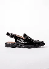 Mocasines tipo slingback de cuero charolado negro, con taco bajo de 1.5 cm y tira frontal decorada con tachas metálicas. Presenta una tira ajustable con hebilla en el talón.