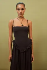 Vestido largo de color negro con escote recto y tirantes finos ajustables. Presenta un diseño con detalle de volante tipo peplum en la cintura y falda con caída fluida.