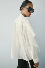 Camisa de hilatura con mezcla de lyocell color blanco, con cuello fluido con pañuelo incorporado, manga larga con puño y bajo redondeado asimétrico. Cierre frontal con botones ocultos.