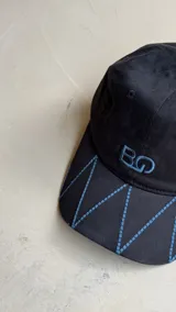 Gorra negra con bordado de las iniciales BG en el frente y diseño de costuras geométricas en la visera.