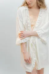 Bata tipo kimono de algodón color blanco, con detalle de volados en el borde frontal y en los puños de las mangas. Presenta un largo por encima de la rodilla y un lazo ajustable en la cintura.