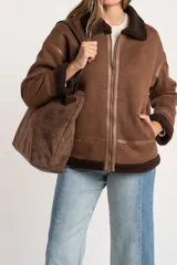 Chaqueta tipo gamulán de calce oversize en color marrón, con cuello y ribetes de corderito en contraste. Presenta cierre frontal de cremallera, bolsillos laterales y detalles de tiras decorativas en material sintético.