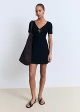 Vestido corto de punto fino en color negro, con escote en pico y detalle de lazo frontal. Presenta mangas cortas y un diseño evasé que aporta movimiento a la prenda.