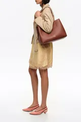 Bolso shopper Bimba Y Lola color marrón, de tamaño grande con exterior de piel vacuna. Presenta doble asa de hombro, cierre con botón de presión y logo grabado en letras doradas en la parte frontal.