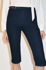 Jeans de tiro medio en denim azul oscuro, con corte capri y pernera entallada. Presenta un diseño con hebilla metálica integrada en la cintura y cierre frontal con cremallera.