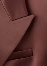 Blazer de corte recto y diseño estructurado, confeccionado en tejido satinado color marrón. Presenta cuello con solapas de muesca, cierre frontal de doble botonadura, bolsillos de solapa en el frente y bolsillo de parche en el pecho. Incluye forro interior y mangas largas con puños abotonados.
