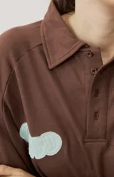 Buzo celeste de manga larga con cuello polo y cierre frontal de botones. Presenta un parche marrón bordado en el pecho.