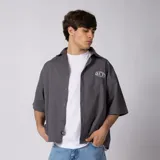 Camisa de manga corta color gris oscuro con corte boxy, confeccionada en 100% algodón de mano suave. Cuenta con cierre frontal de botones, cuello clásico y un pequeño bordado en el pecho.