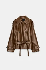 Trench corto de efecto piel color marrón, con cuello de solapa, cierre frontal cruzado con botones y cinturón lazada del mismo material. Presenta mangas largas con trabillas en hombros y puños, y bolsillos de vivo en el delantero.