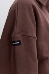 Sudadera de algodón color marrón, con cuello polo y cierre frontal de botones. Presenta un diseño holgado, mangas largas con puños elásticos y un pequeño parche bordado en forma de corazón en el pecho.
