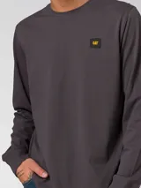 Remera de manga larga color gris oscuro, con cuello redondo y puños acanalados. Presenta un pequeño parche rectangular con el logo de la marca en el lado izquierdo del pecho.
