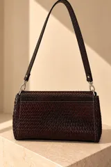 Cartera tipo bandolera de cuero vacuno con textura trenzada en tonos marrones y negros. Presenta una solapa frontal con cierre magnético y una placa metálica dorada con el logo de la marca. Incluye una correa larga y ajustable con herrajes plateados.