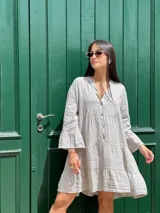 Vestido corto de lino color gris claro, con escote en V y botones de coco en el frente. Presenta mangas largas con volados acampanados y un diseño amplio con frunces en el torso y falda.