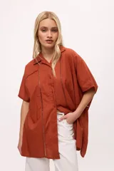 Camisa de manga corta y corte oversize en color tostado, confeccionada en algodón. Presenta cuello clásico, cierre frontal con botones y detalles de calados verticales en el frente y mangas.